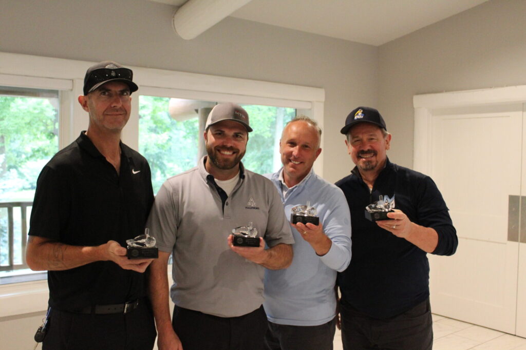 Four men holding trophies indoors smiling.